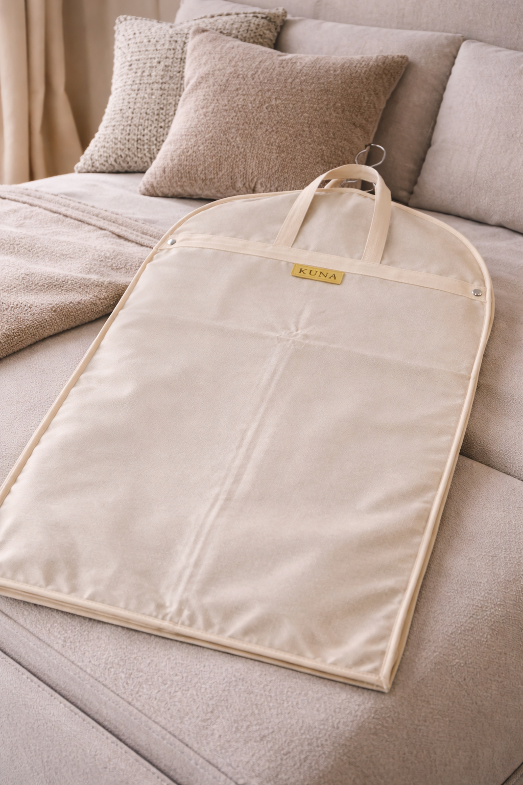 Beige garment bag with a brand logo on a bed with pillows