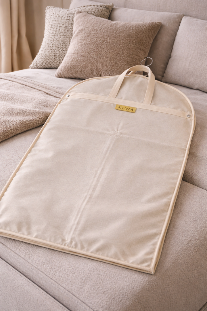 Beige garment bag with a brand logo on a bed with pillows